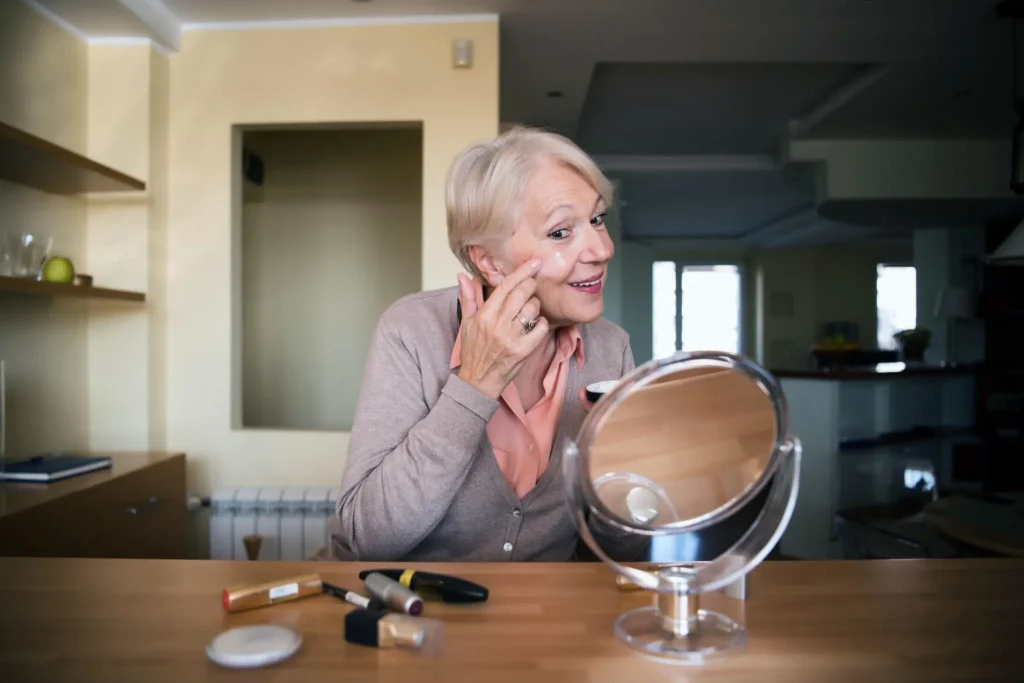 Mature woman applying estradiol cream to her face while looking in a mirror as part of an anti-aging skincare routine.