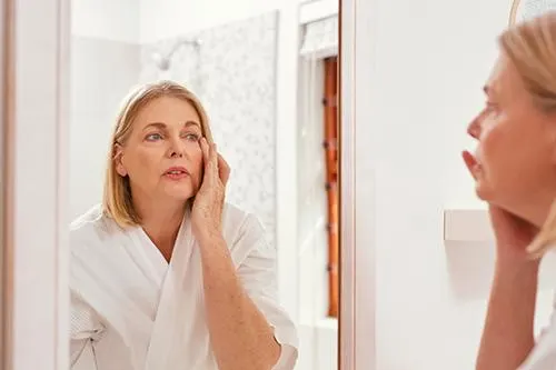 Mature woman examining facial skin changes in a mirror during menopause.