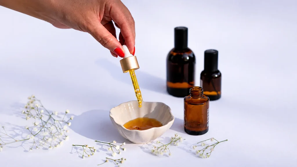 Hand holding a dropper with natural facial serum above a bowl, surrounded by amber skincare bottles and flowers in a minimalist beauty setup.