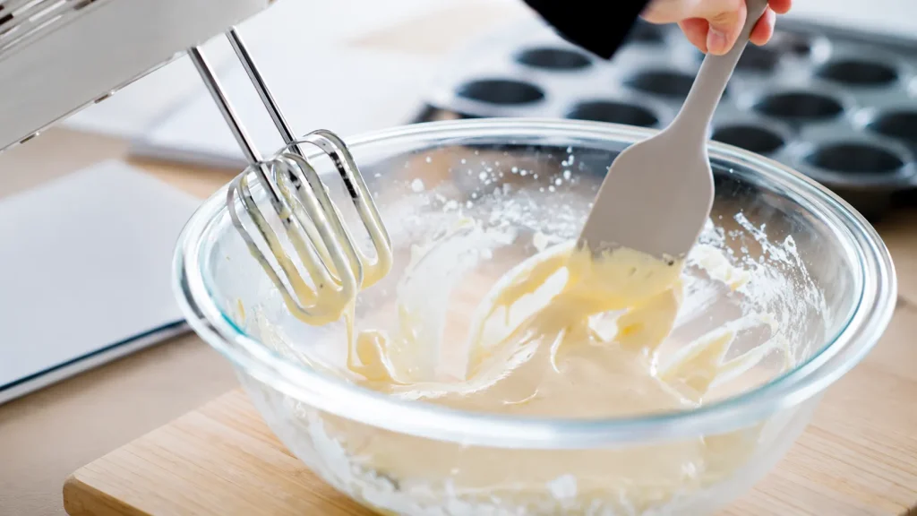 Hand mixing creamy whipped mixture in a glass bowl with an electric hand mixer on a kitchen countertop.